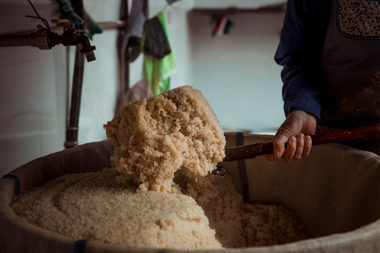 about-img A person kneads dough in a large basin with a wooden spoon in a rustic kitchen setting.