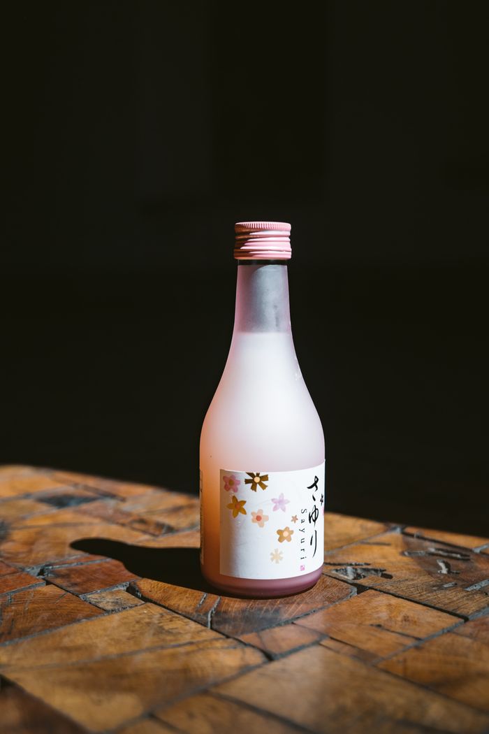 contact-img Pink capped sake bottle with floral label on a wooden surface, highlighted by natural light.